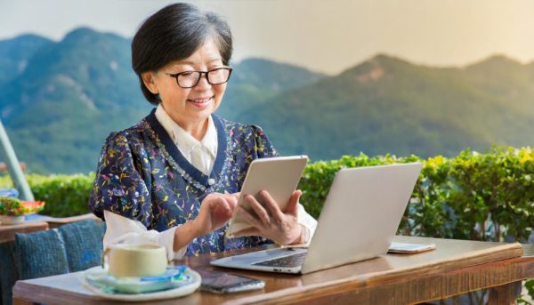 For illustration purposes only. An AI-generated image by Adobe Firefly of an older female adult of Korean heritage using a tablet; other portable tech devices nearby on the table include a laptop and smart phone. 